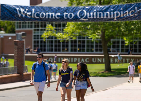Three students walk and talk under a Welcome to Quinnipiac! sign on a sunny day