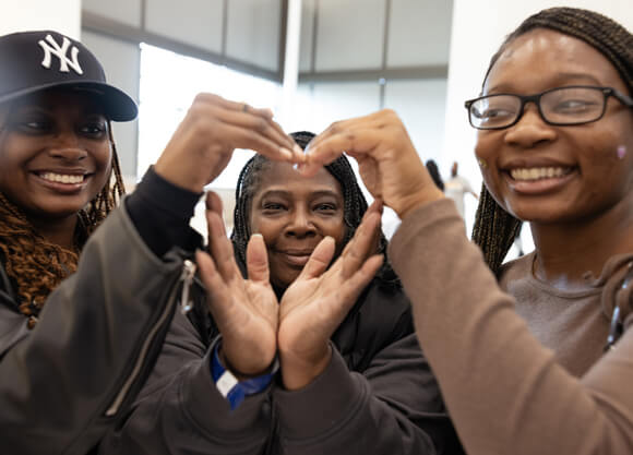 Three students of color make their hands into a heart and smile at a multicultural reception