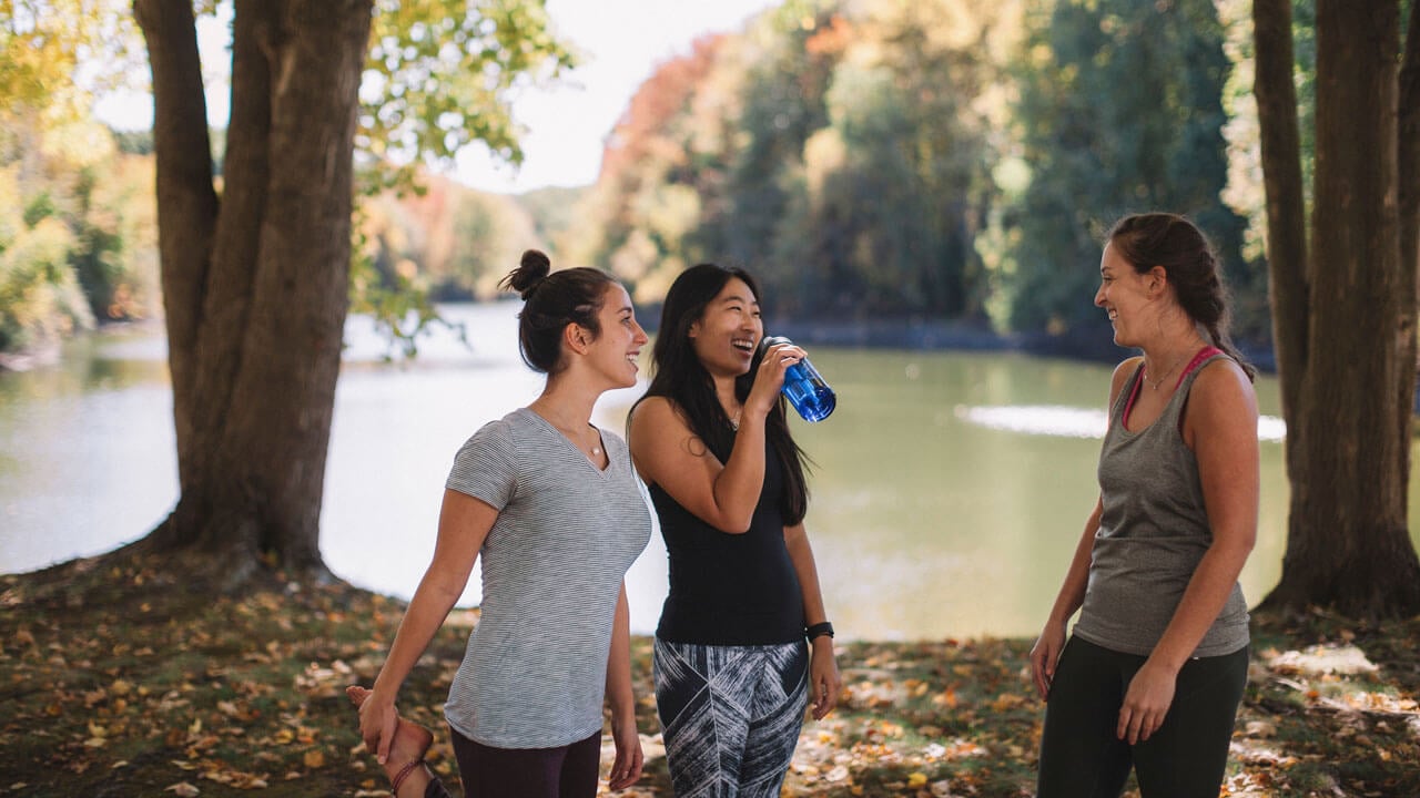 Students in workout gear standing outside by lake and are laughing