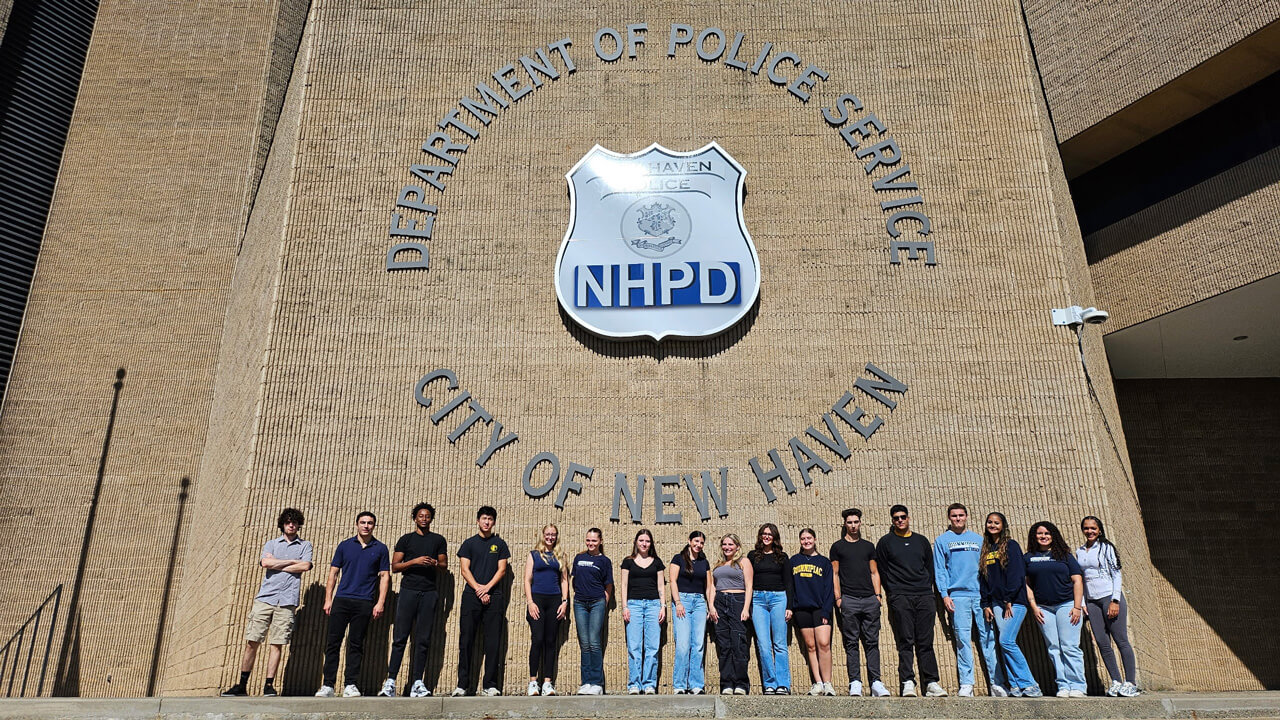 A group of 17 students stands outside the New Haven Police Department building, underneath the logo on the wall, at the top of a staircase.