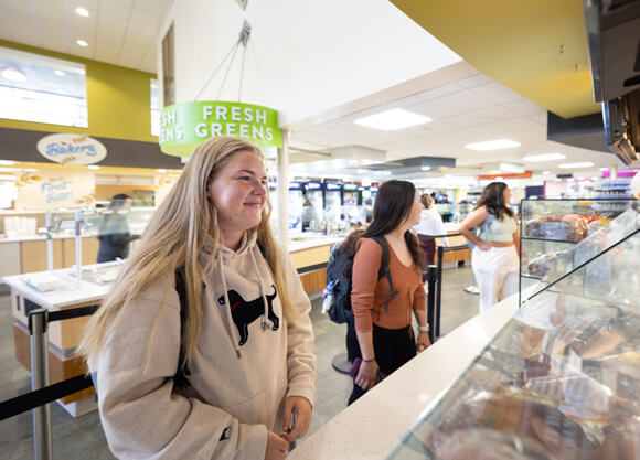 A student waits for food at a dining station in the Cafe Q with a Fresh Greens sign behind her
