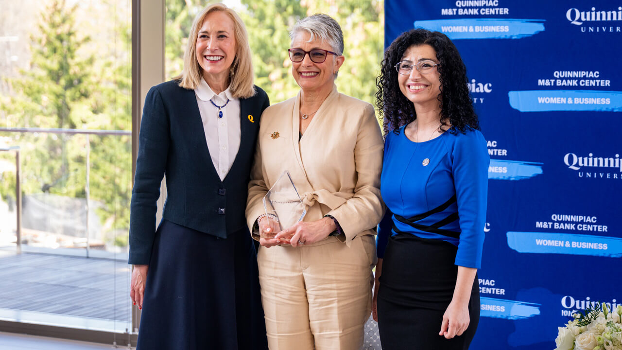 President Hardin, Fran Pastore, and Tuvana Rua pose smiling in front of a blue Quinnipiac banner as Pastore holds her glass award.