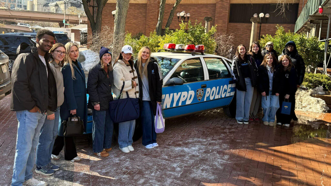 A group of 12 students and faculty members stand on either side of an NYPD police car.