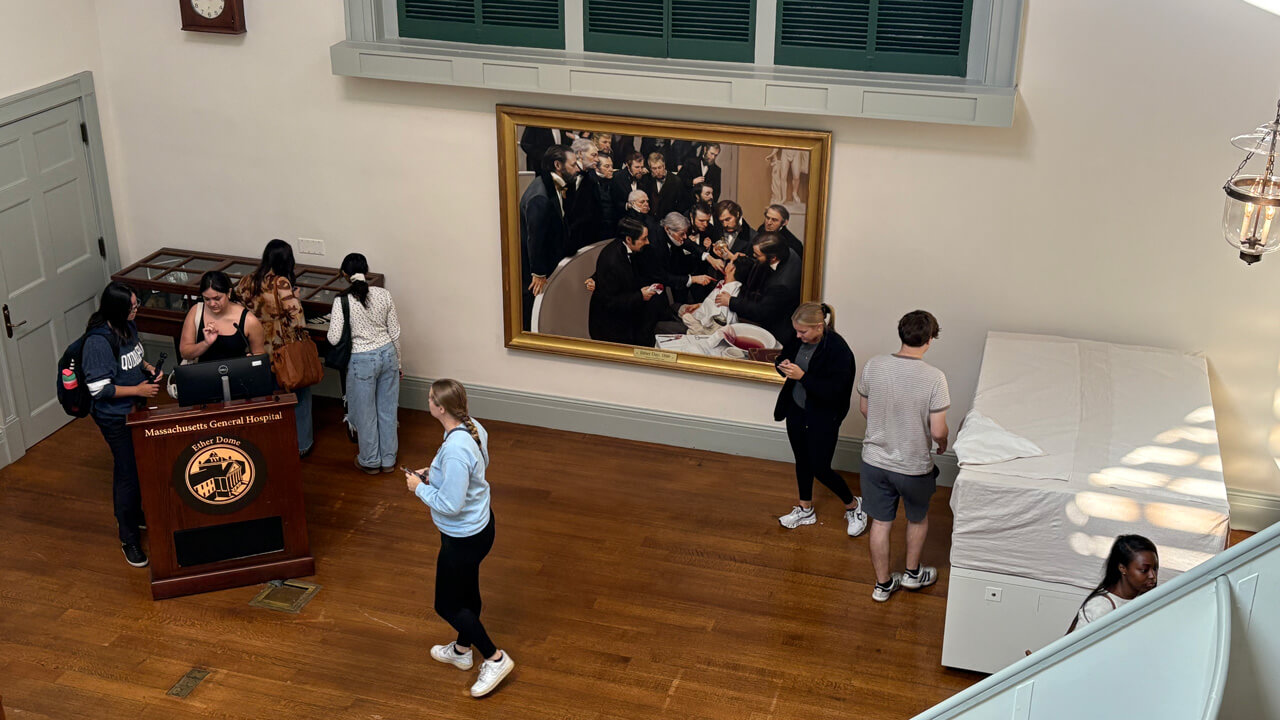 An overhead shot of students looking at various medical exhibit displays, including a podium with a computer on it and a painting of a surgery.