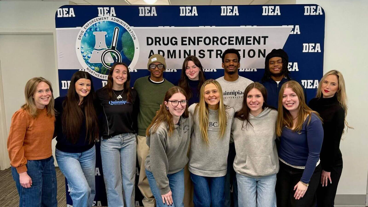 A group of 12 students and faculty members stands in front of a Drug Enforcement Administration backdrop with a graphic of a beaker and a magnifying glass.
