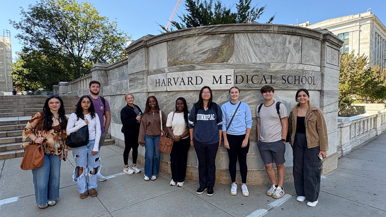 A group of 10 students stands in front of a stairwell and a stone Harvard Medical School sign.