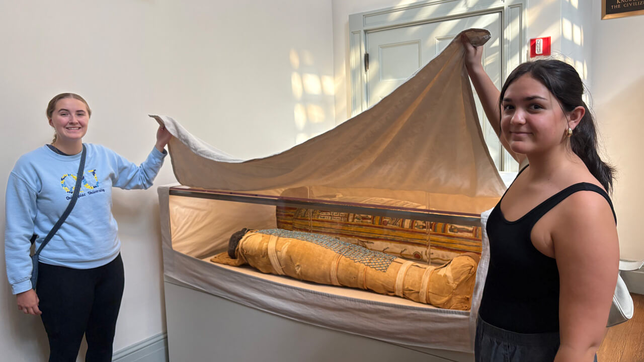 Two female students lift a shroud over a glass case sarcophagus display.