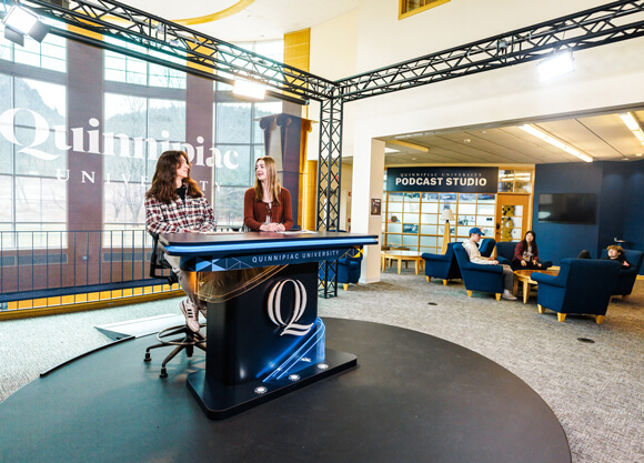 Two students sit at the broadcast desk of the Quinnipiac open air studio
