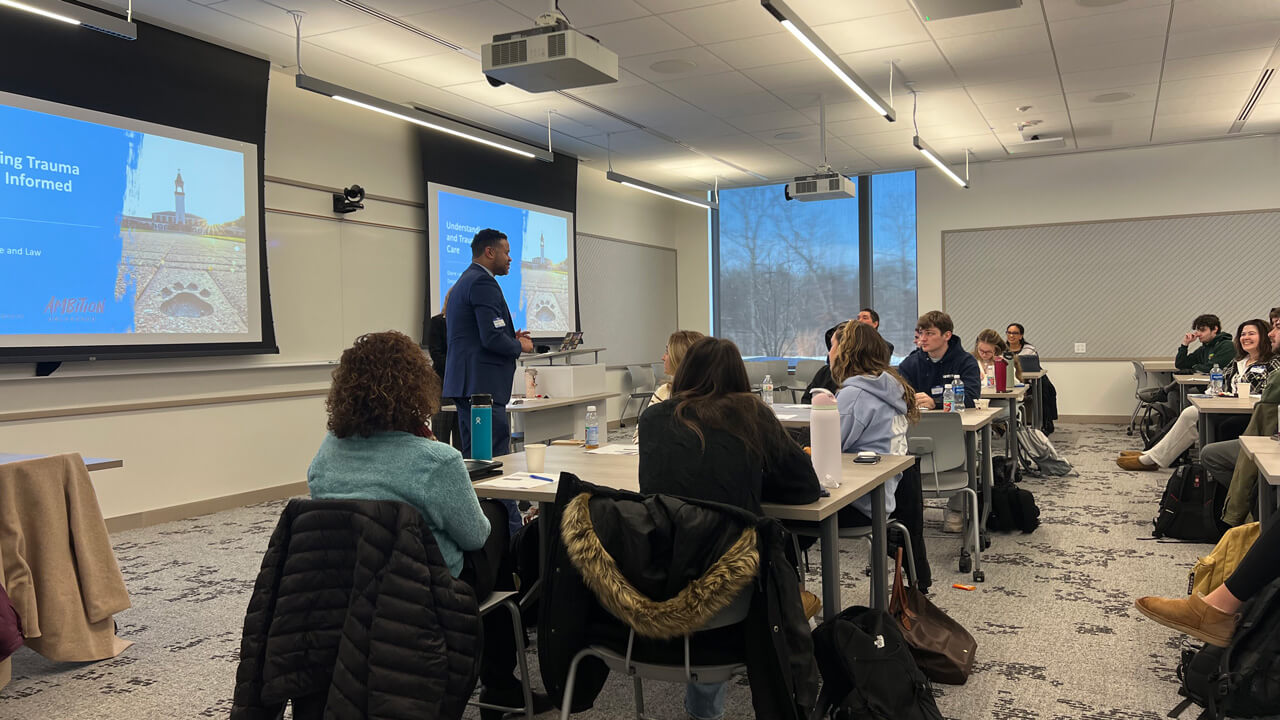 A speaker speaks in front of the class with a PowerPoint slide reading "Understanding Trauma and Trauma Informed Care."
