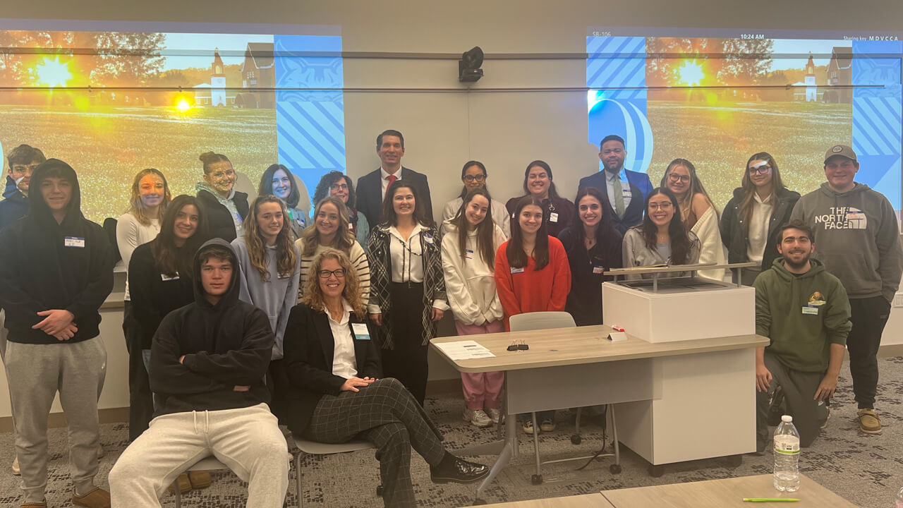 A group of 20 students, plus speakers and faculty, pose in a group in front of the classroom in front of two projector screens.
