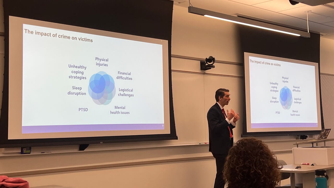 Marc Pelka speaks in front of the classroom next to a PowerPoint slide reading "The impact of crime on victims," with smaller bubbles that say “physical injuries,” “financial difficulties,” “logistical challenges,” “mental health issues,” “PTSD,” “sleep disruption” and “unhealthy coping strategies.”