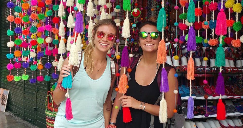 Two students stand outside a market selling colorful fabric in Peru