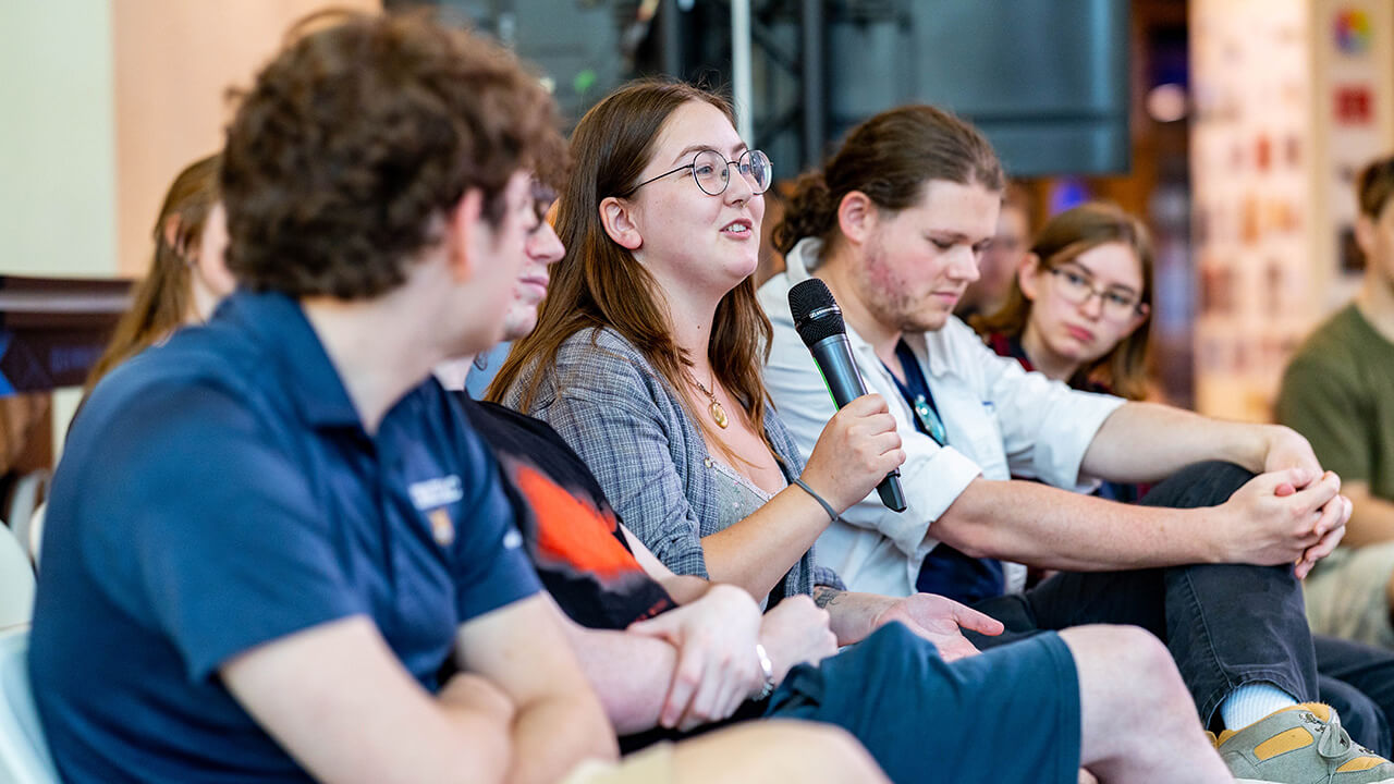 A female student speaking into a microphone while sitting in a crowd