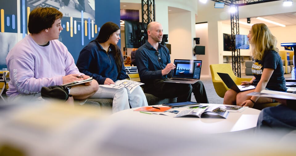 A professor demonstrates information on his laptop to three students outside the podcast and open air studios