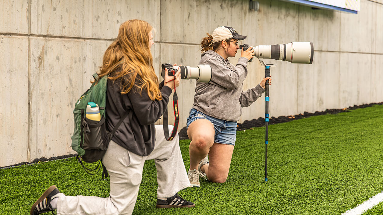 2 female student kneeling with large sports cameras taking photos