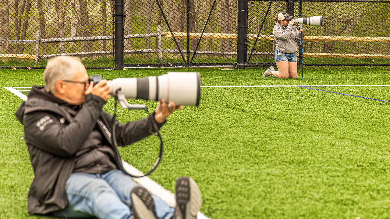 Simon Bruty sitting in the grass with a large sports camera in the foreground with a female student kneeling with a large sports camera in the background