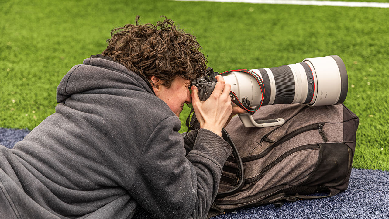 A male student laying in the grass resting a large sports camera on a backpack