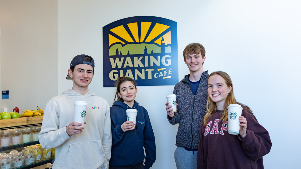 Two male students and two female students hold up Starbucks coffee cups in front of the Waking Giant Café logo.