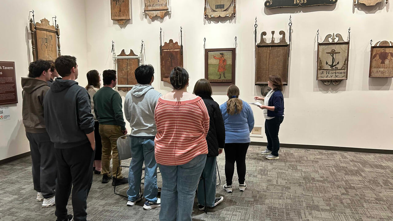 A photo shot from behind of nine students looking at a wall of framed Revolutionary War images and symbols, with a female tour guide giving a presentation.