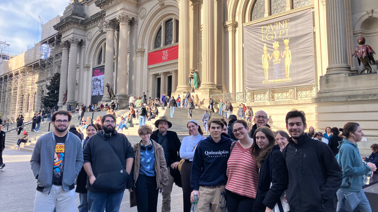 A group of 13 students and faculty members stands in front of the Metropolitan Museum of Art, below banners reading "The Met" and "Divine Egypt."