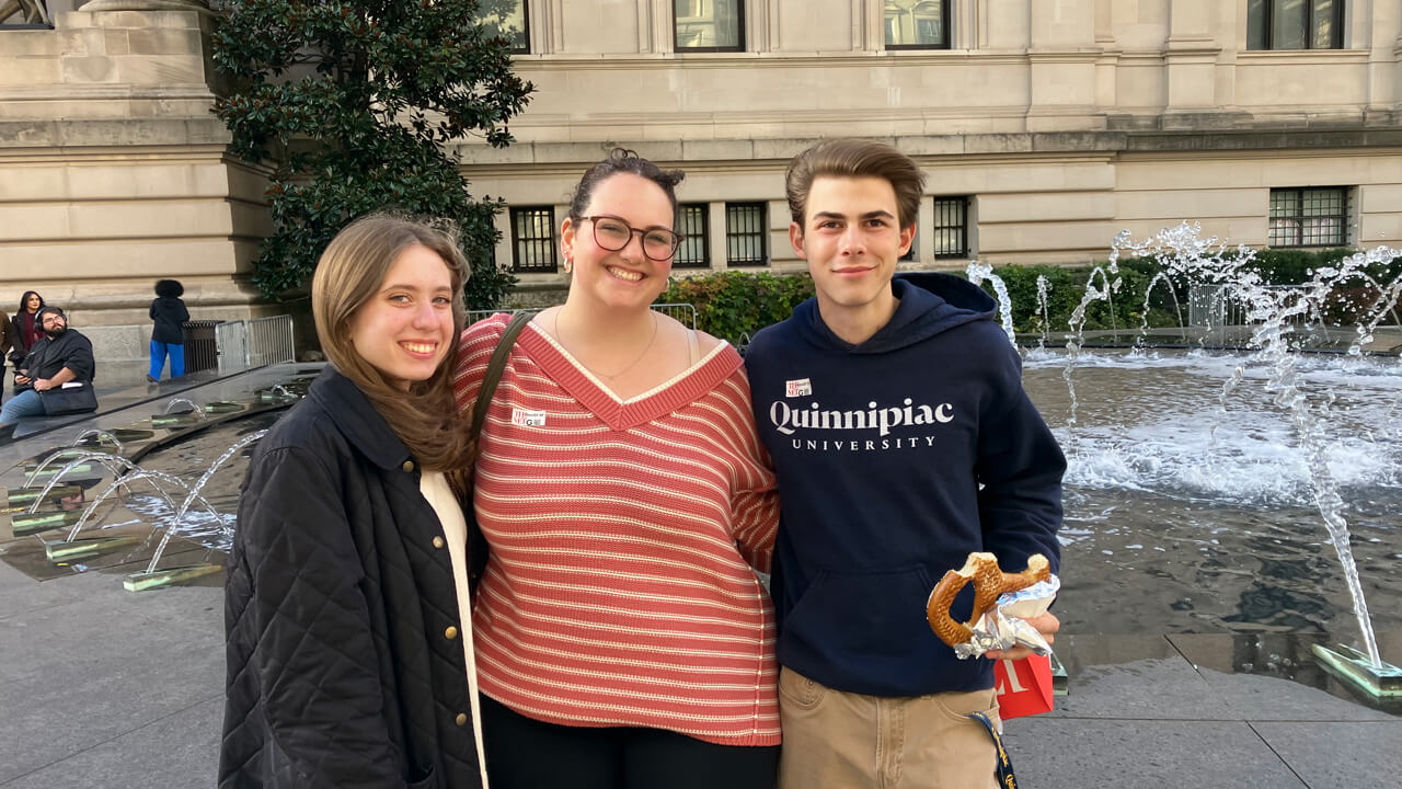 Three students stand smiling in front of a fountain outside the Met, one holding a New York soft pretzel and a Met shopping bag.