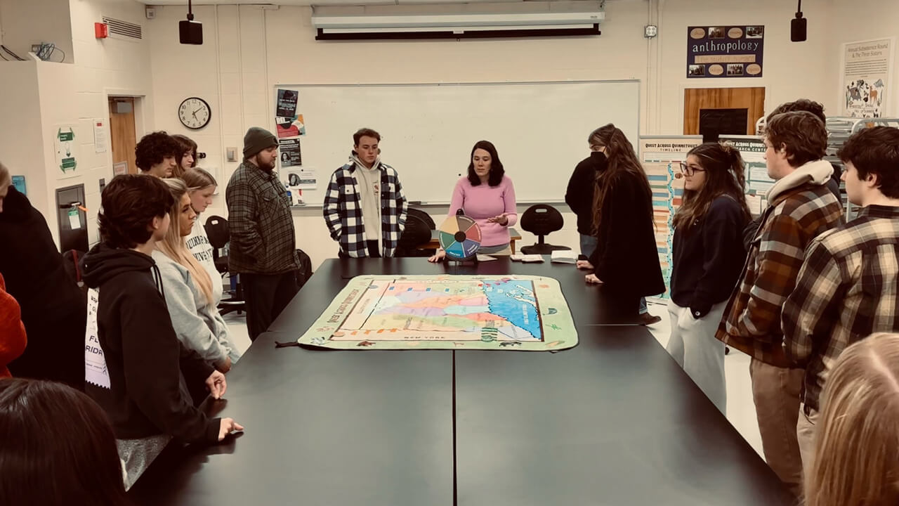 A professor and group of students grouped around a table with a colorful Connecticut map on it. A poster on the classroom wall reads "Anthropology."