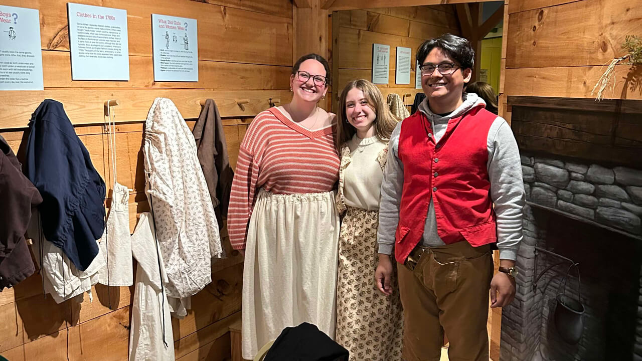 Three students pose in a wartime home exhibit, trying on 1700s-era costumes.