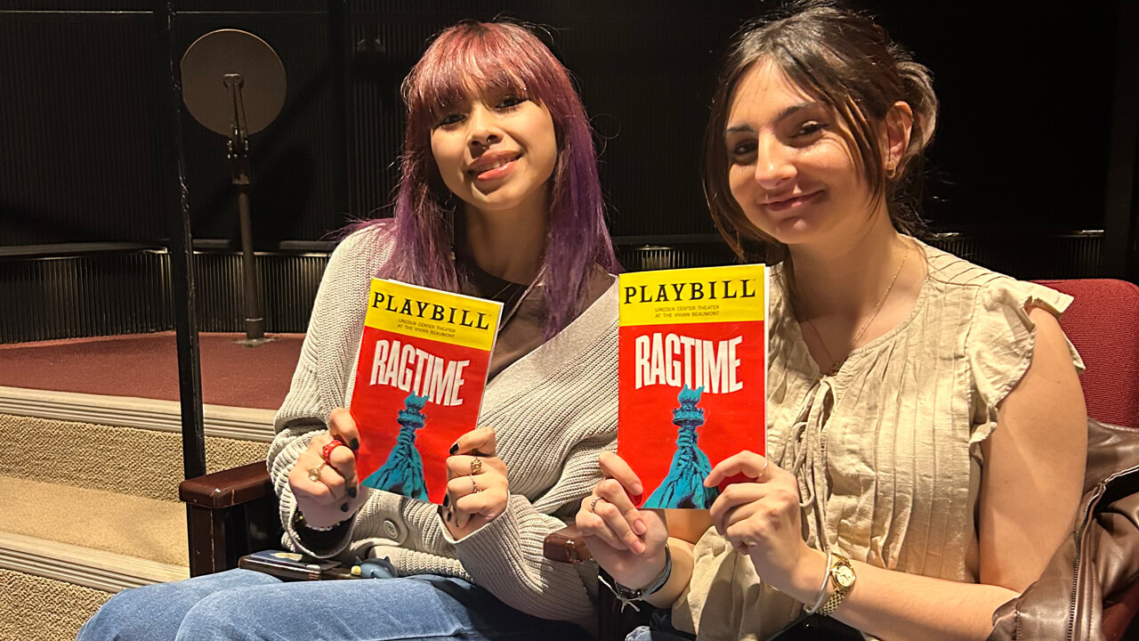 Two female students sitting in their theatre seats holding up their playbills.