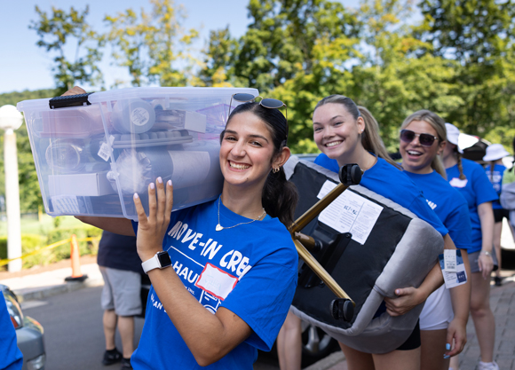 Three students in blue shirts carry bins and furniture during move in