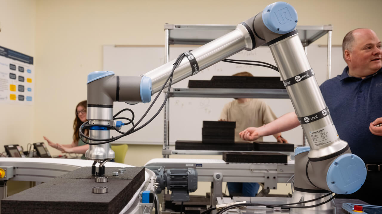 A sideways shot of a robot arm and other equipment with a professor and students in the background.