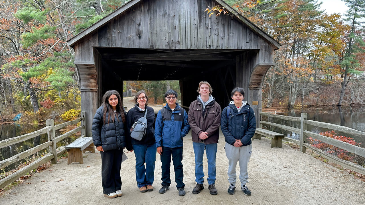 A group of five participants standing in front of a covered bridge surrounded by fall foliage.