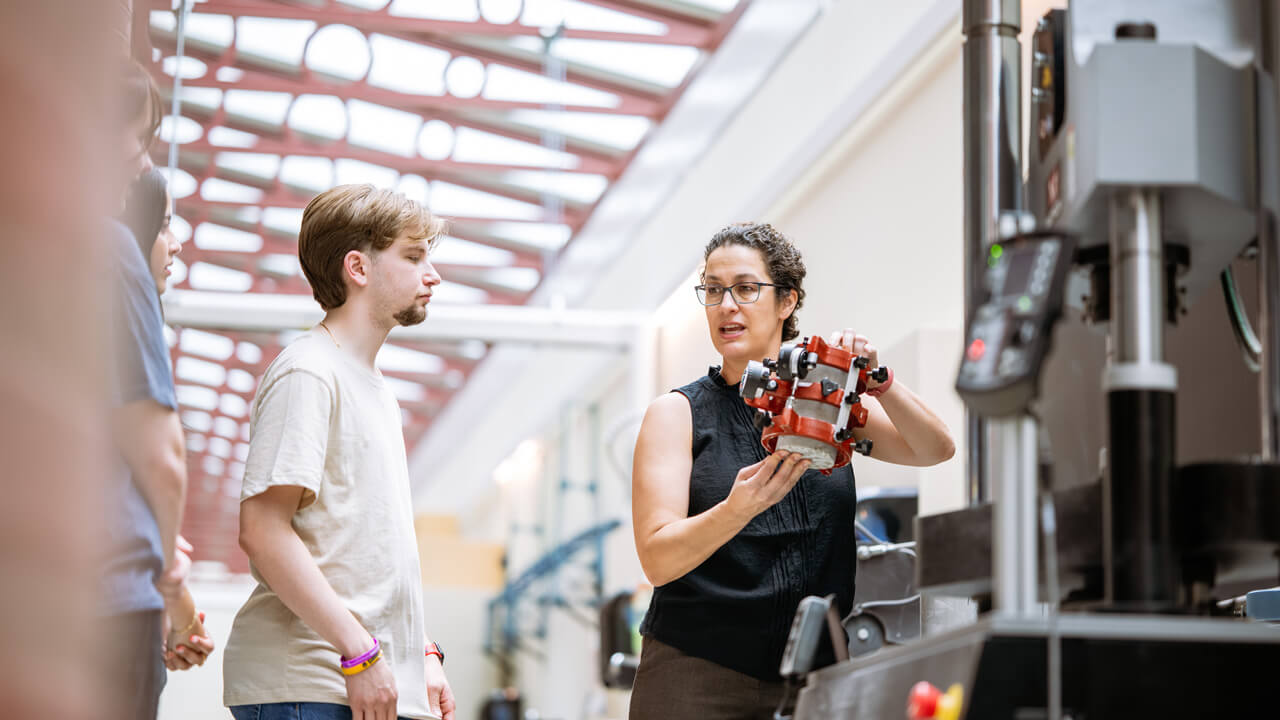 Students look on while a female faculty member holds up a piece of equipment while speaking.