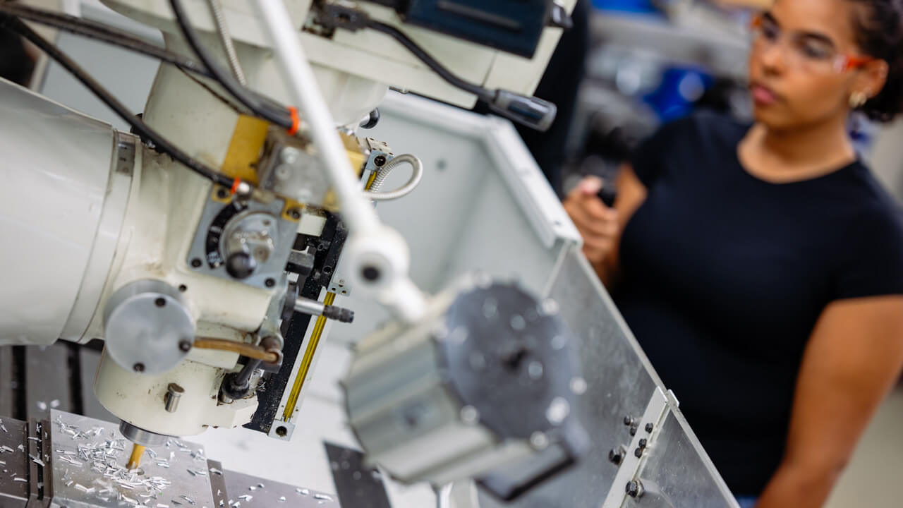 A professor pointing to a screen on a piece of machinery while a student looks on. The machinery is labeled "PROGRAM SPINDLE CONTROL."