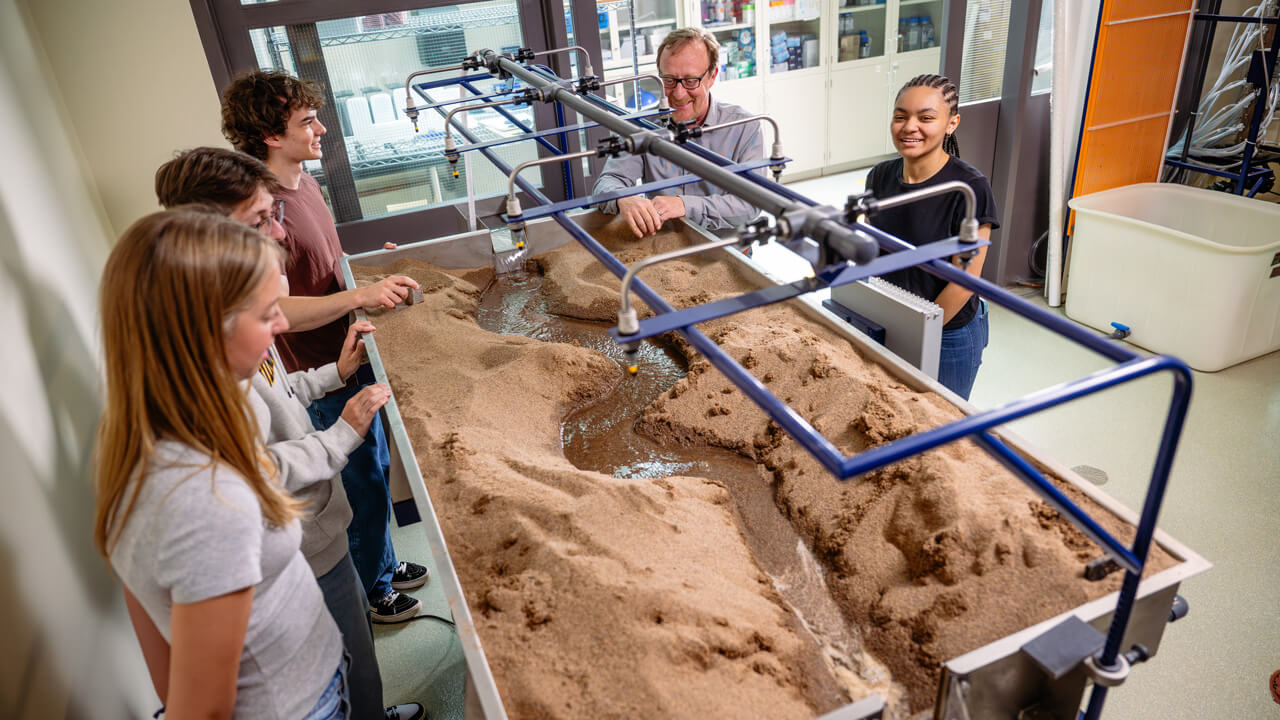 A shot from above of four students and a faculty member looking into a basin in the Fluids Lab filled with soil and water.
