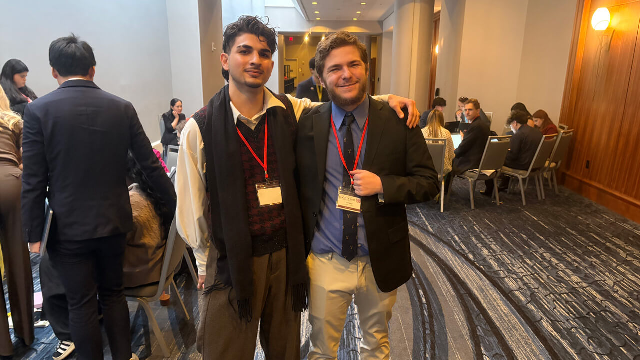 Two male students with name badges smiling with their arms around each other, in a busy conference room.