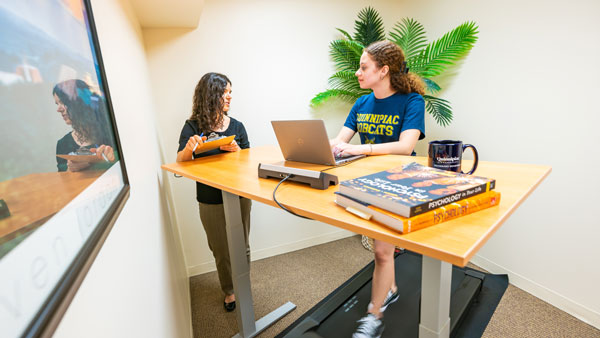 A female student working at a standing desk while on a walking pad being studied by a woman with a clipboard