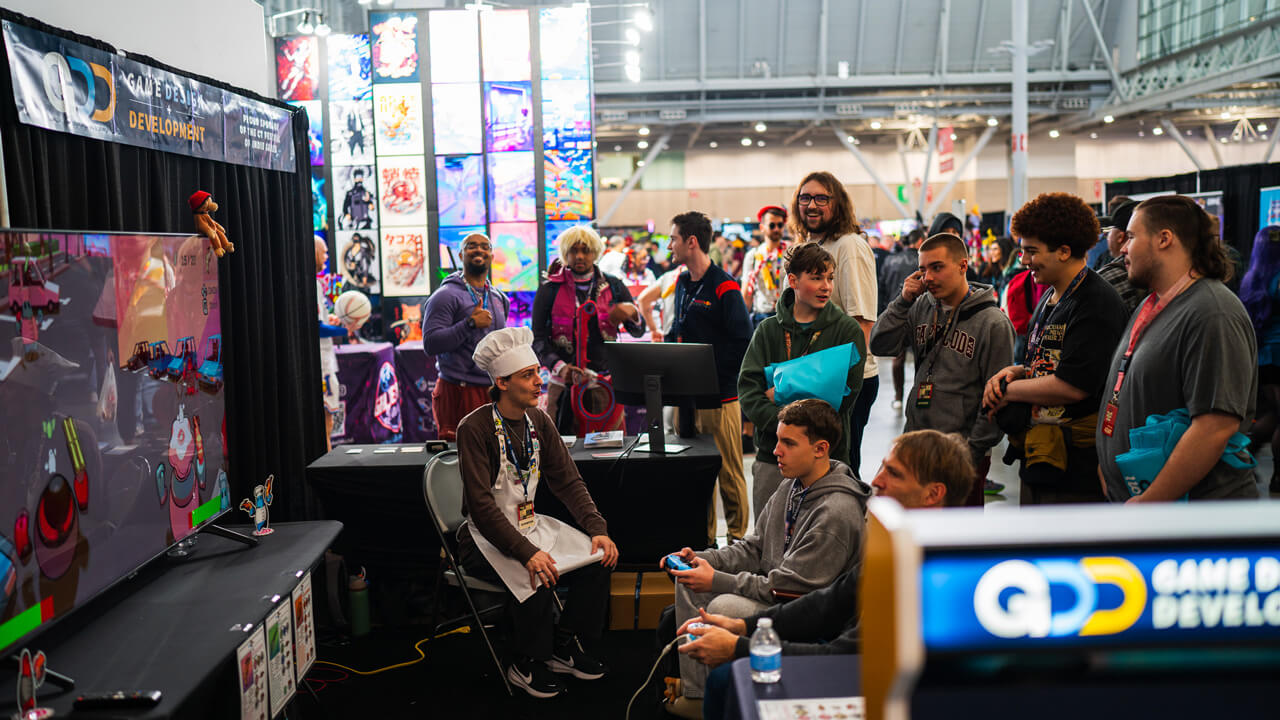 Participants around the Quinnipiac Pax East booth, watching two players gaming on the monitor, with the event crowd in the background.