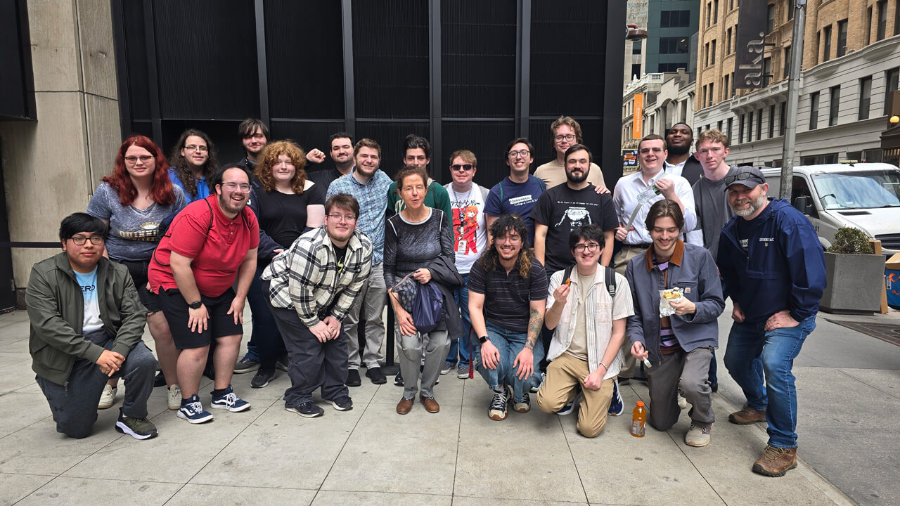 Twenty-two students and faculty members smiling in a group outside the T2 building.