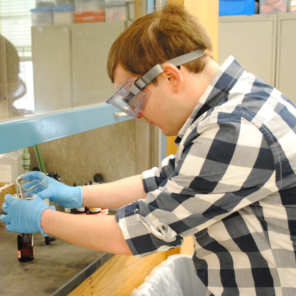 Mark Elsesser in lab goggles and a checkered shirt, pouring a beaker into a funnel.