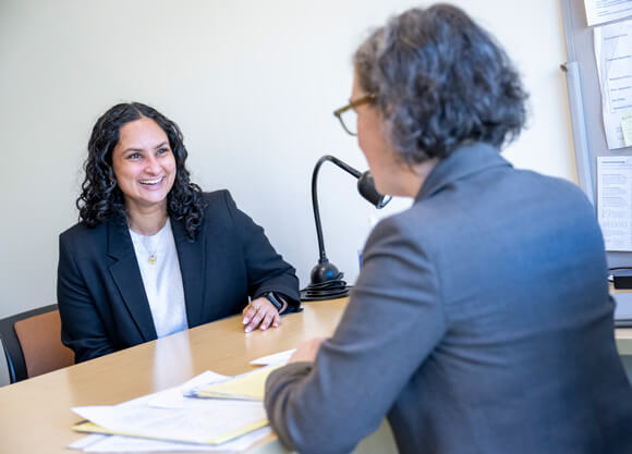 A female grad student sitting at the desk of a professor, talking to the professor