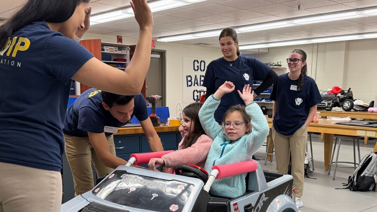A photo of a young girl in a toy car with her hands in the air and a female student in front of her with her hands up.