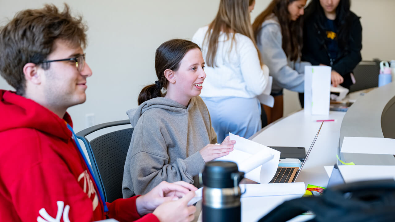 Two students smiling and chatting while folding pieces of paper. Another group of three students stands in the background.