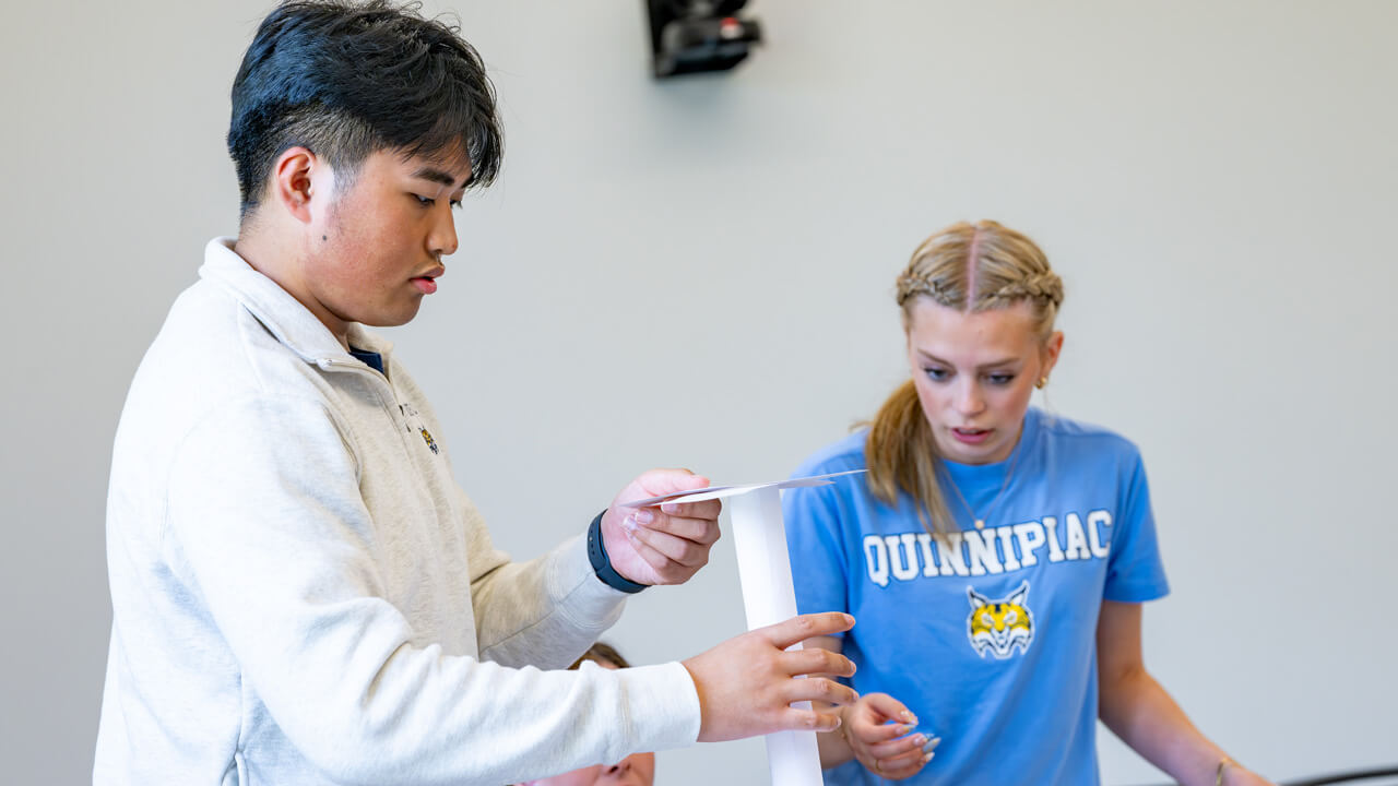 Two students standing at a desk and carefully balancing pieces of paper for an activity.