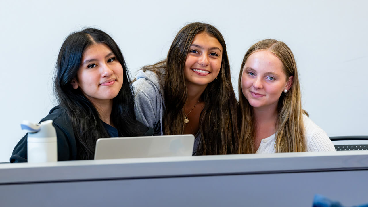 Three female students smiling at the camera while crowded around a desk.