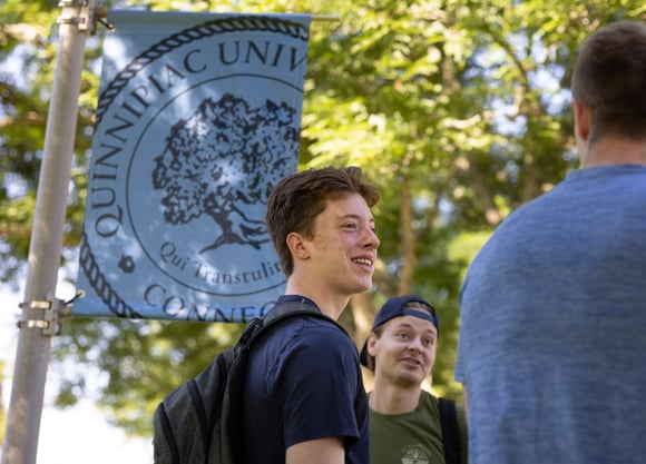 A transfer student smiles under a banner with the Quinnipiac seal outside on a sunny day