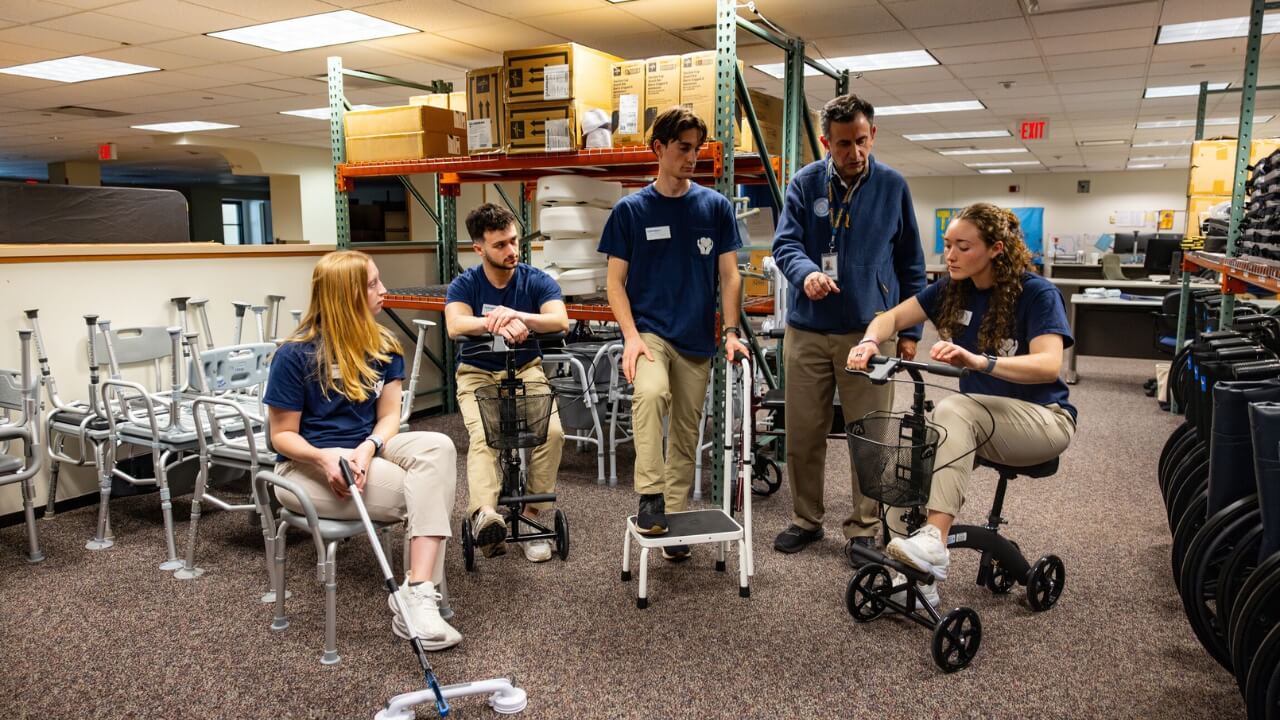 Volunteers and a staff member review and demonstrate mobility devices inside a building. Walkers, rollators, shower chairs, wheelchairs, and other assistive equipment are arranged throughout the room.