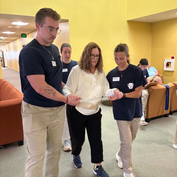 Two staff members assist a participant during a mobility exercise in a brightly lit rehabilitation setting. The participant walks between them while each staff member provides support, with other people seated and observing in the background.