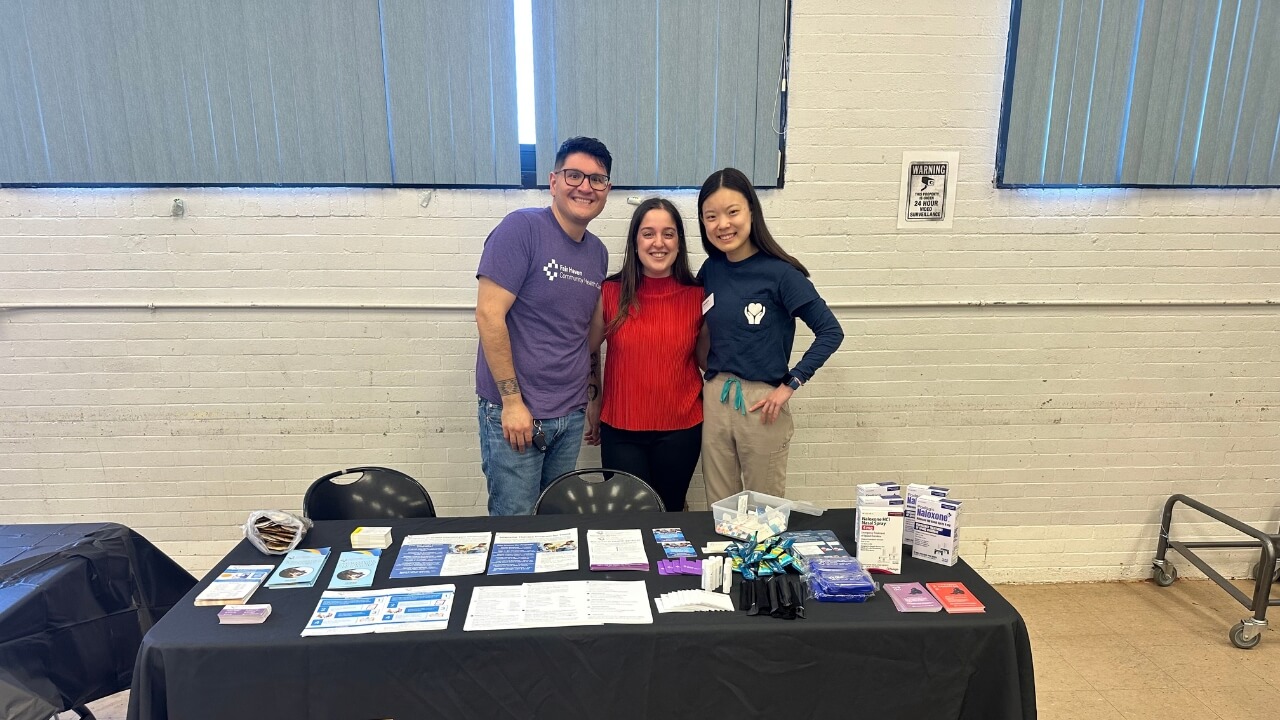 Three volunteers stand behind an informational display table at a community event. The table is covered with educational materials, harm reduction supplies, and naloxone kits arranged for attendees.
