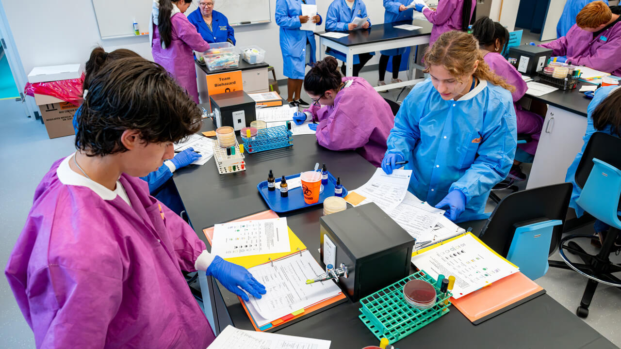 An overhead shot of a lab table with students writing and working, dressed in bright purple and blue scrubs.