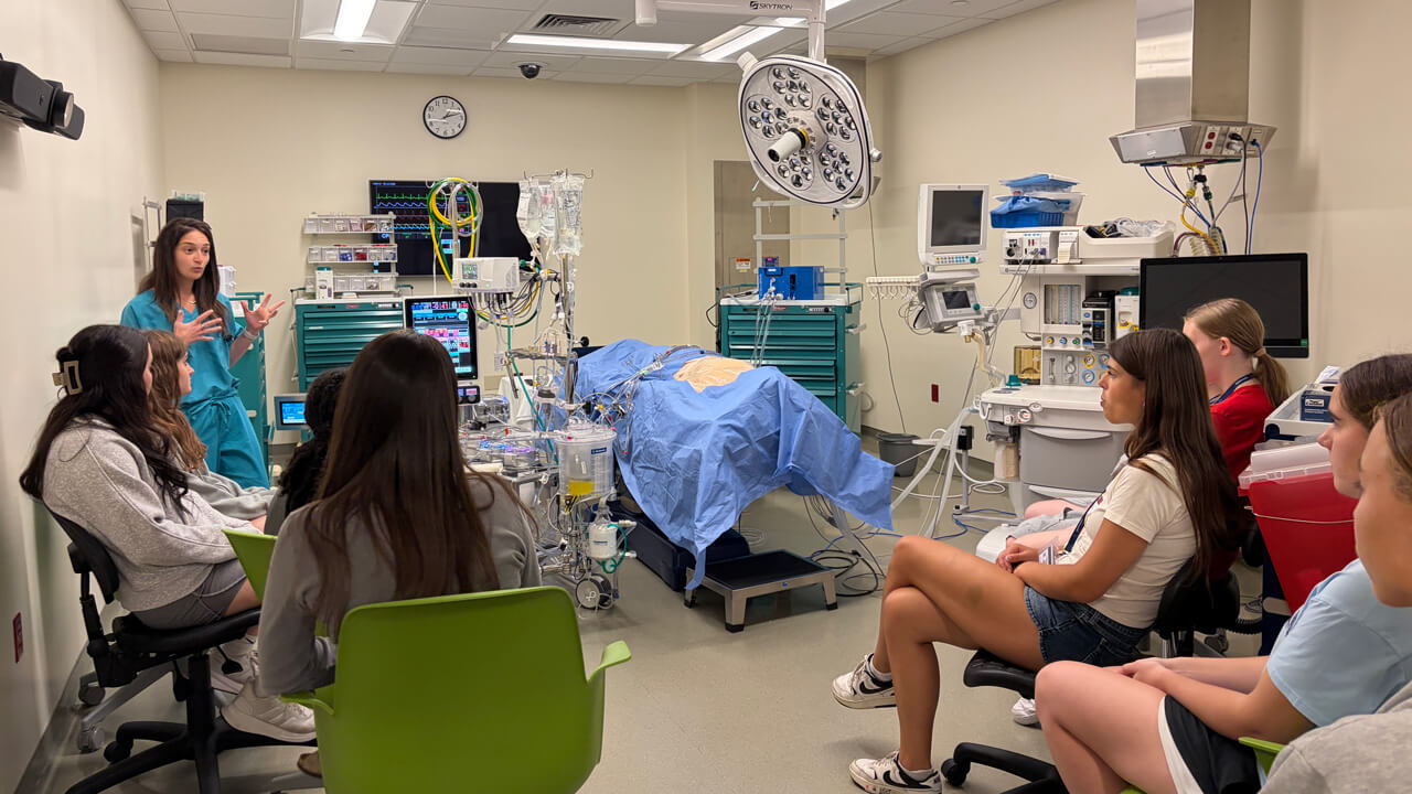 A woman in green scrubs talks to a group of student participants next to a hospital bed simulation.